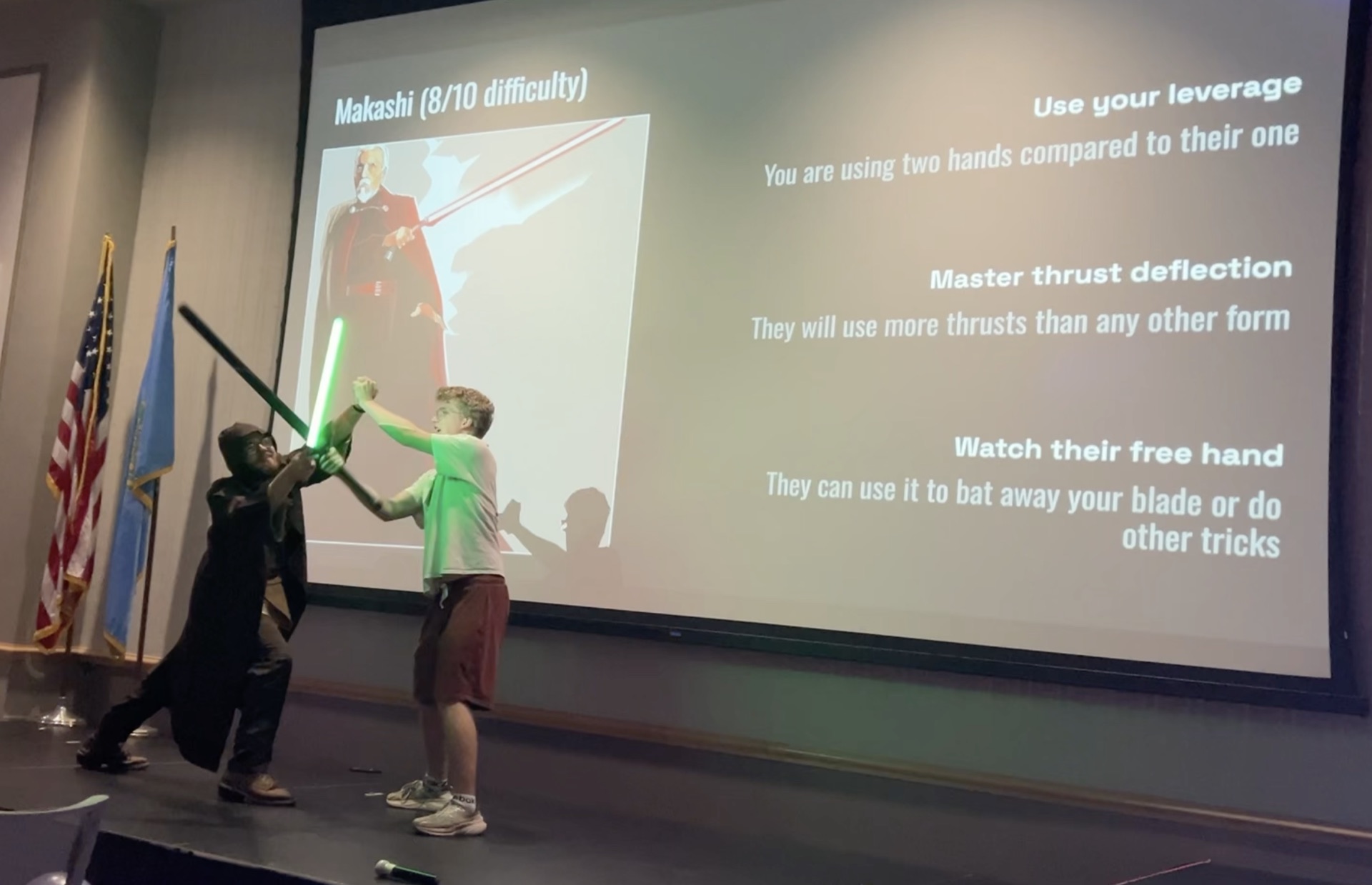 A lightsaber demonstration on stage at Multi-Club Mashup IV, with a slide of sword-fighting tips on the screen behind