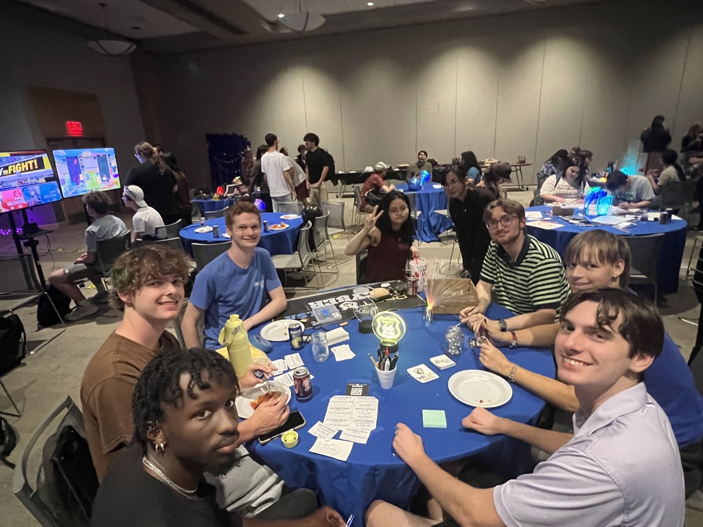 A full table of students smiling at the camera, with games on screens and busy tables behind them